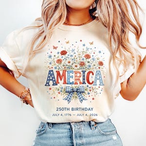 May include: Cream-colored t-shirt with the word "AMERICA" in red, white, and blue letters, surrounded by a floral design. The shirt also features the text "250TH BIRTHDAY JULY 4, 1776 - JULY 4, 2026".
