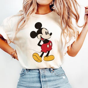 May include: Cream-colored t-shirt featuring a classic Mickey Mouse graphic. The cartoon character is depicted in black, red, and yellow, with a vintage aesthetic. The shirt is paired with blue jeans.