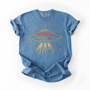 May include: A blue short-sleeve t-shirt featuring a graphic print of a flying saucer emitting a yellow light beam. The saucer is depicted in shades of red, green, and blue, surrounded by yellow star-like shapes against the blue fabric background.