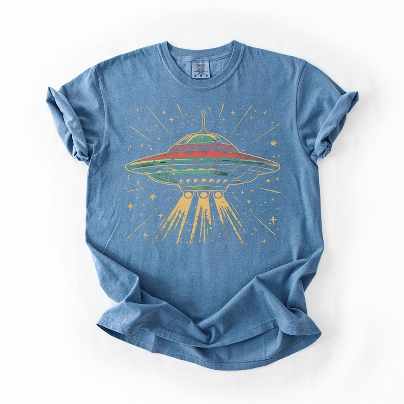May include: A blue short-sleeve t-shirt featuring a graphic print of a flying saucer emitting a yellow light beam. The saucer is depicted in shades of red, green, and blue, surrounded by yellow star-like shapes against the blue fabric background.