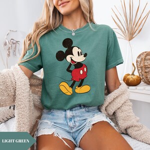 May include: Light green t-shirt featuring a vintage-style Mickey Mouse graphic. The cartoon character is depicted in his classic outfit: red shorts, yellow shoes, and black ears. The shirt has a slightly faded appearance, giving it a retro aesthetic. The text "LIGHT GREEN" is at the bottom.