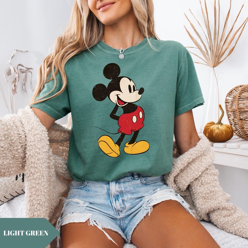 May include: Light green t-shirt featuring a vintage-style Mickey Mouse graphic. The cartoon character is depicted in his classic outfit: red shorts, yellow shoes, and black ears. The shirt has a slightly faded appearance, giving it a retro aesthetic. The text "LIGHT GREEN" is at the bottom.