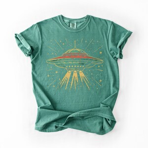 May include: A teal green short-sleeve t-shirt featuring a graphic print of a flying saucer emitting a light beam. The saucer is depicted in shades of red and yellow with radiating lines and star shapes surrounding it on the front of the garment.