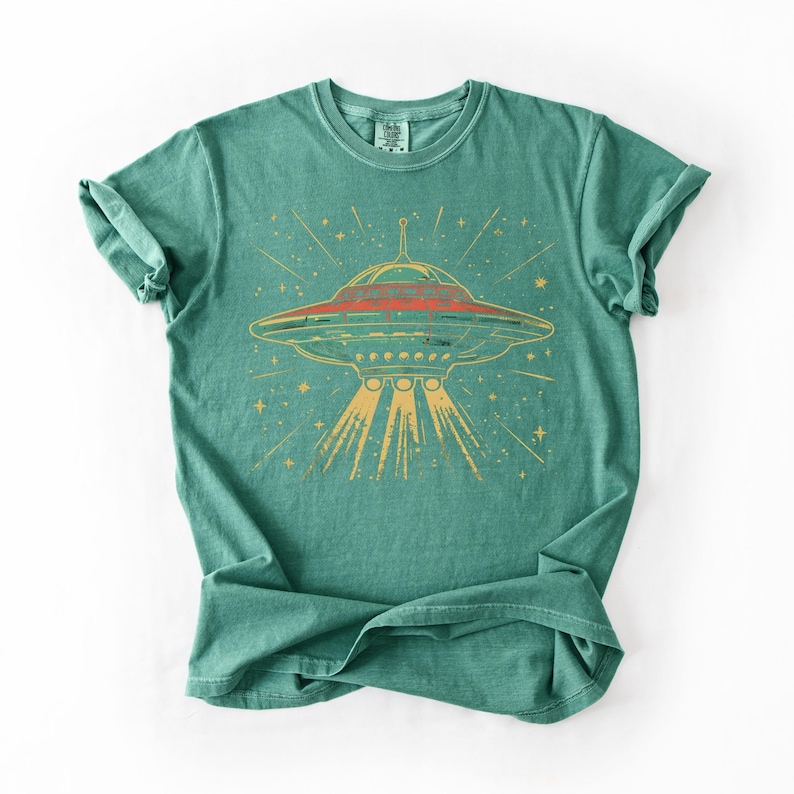 May include: A teal green short-sleeve t-shirt featuring a graphic print of a flying saucer emitting a light beam. The saucer is depicted in shades of red and yellow with radiating lines and star shapes surrounding it on the front of the garment.