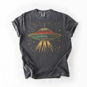 May include: A charcoal gray short-sleeve t-shirt featuring a graphic print of a flying saucer emitting a yellow light beam. The saucer is depicted in shades of green and red against a background of yellow stars and lines on the dark fabric.