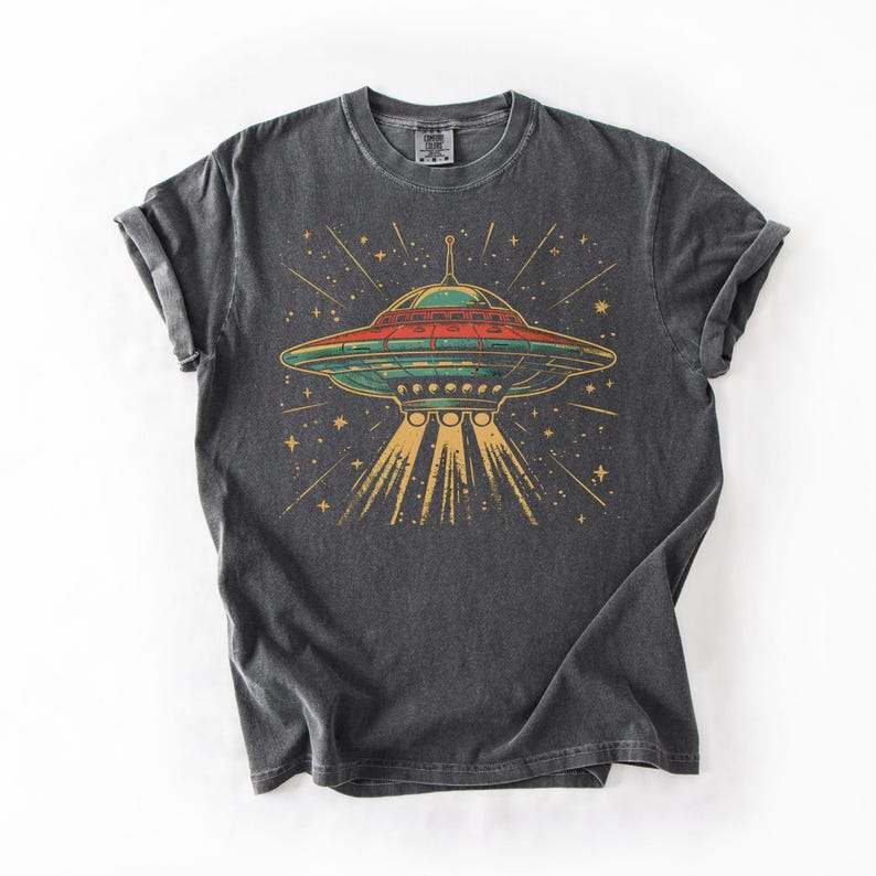 May include: A charcoal gray short-sleeve t-shirt featuring a graphic print of a flying saucer emitting a yellow light beam. The saucer is depicted in shades of green and red against a background of yellow stars and lines on the dark fabric.