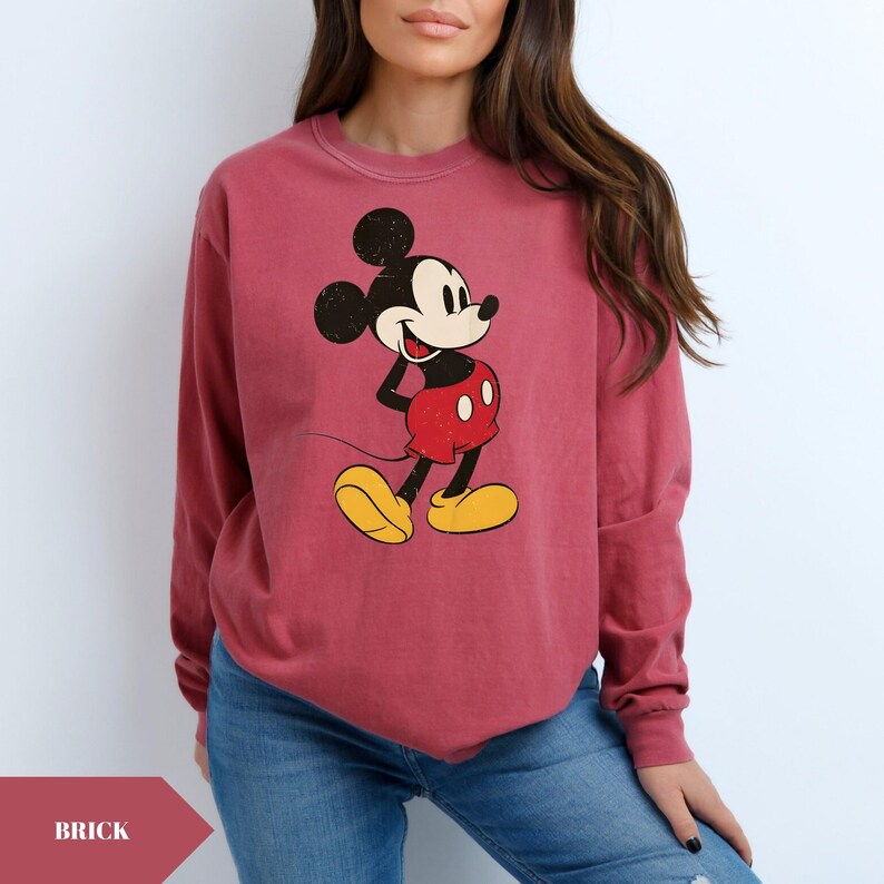 May include: Brick-colored long-sleeve sweatshirt featuring a distressed graphic of Mickey Mouse in his classic pose. Mickey is depicted in black, red, and yellow. The sweatshirt is made of a soft material and has a crew neck.