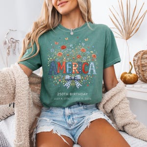 May include: A teal t-shirt with the word "AMERICA" in red, white, and blue letters, surrounded by a floral design. The shirt also features the text "250TH BIRTHDAY JULY 4, 1776 - JULY 4, 2026".