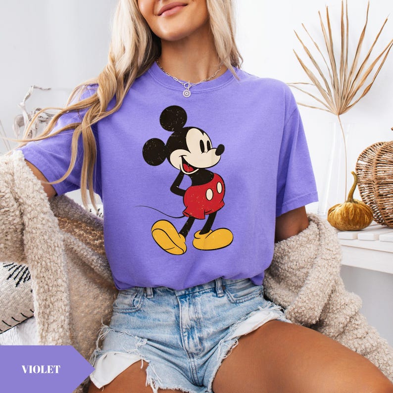 May include: Violet-colored t-shirt featuring a classic Mickey Mouse graphic. The cartoon character is depicted in his iconic red shorts, yellow shoes, and black gloves. The shirt is a comfortable, casual style, perfect for everyday wear.