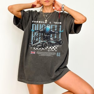 May include: A charcoal gray oversized t-shirt featuring a graphic of a Formula 1 race car and a driver in a blue helmet. The text reads Formula 1 Team, Russell, and 63, accompanied by a small British flag and a checkered pattern design.