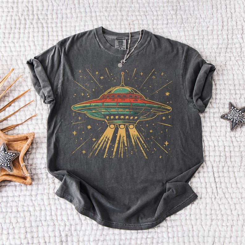 May include: Dark gray t-shirt featuring a colorful illustration of a retro UFO with a red top, green midsection, and gold beams. The design includes stars and lines, creating a space-themed graphic. The shirt is styled with rolled-up sleeves.