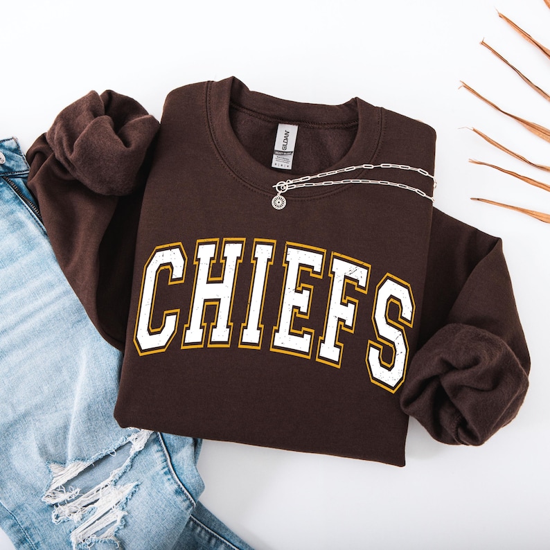 May include: A brown sweatshirt with the word "CHIEFS" in white block letters with gold trim. A silver necklace with a pendant is draped over the sweatshirt. Light blue denim jeans are visible in the background.