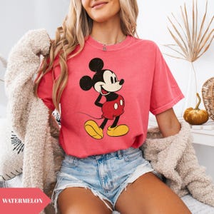 May include: A watermelon-colored t-shirt featuring a classic Mickey Mouse graphic. The cartoon character is depicted in black, red, yellow, and white. The word "WATERMELON" is printed on a pink banner at the bottom left of the image.