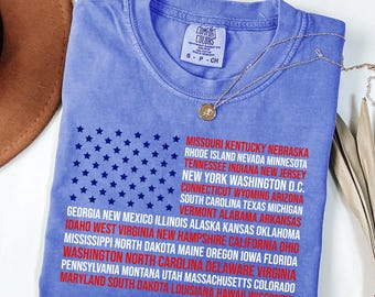 May include: A blue Comfort Colors t-shirt featuring a graphic of the American flag made of stars and the names of the states. A gold necklace with a pendant is draped across the shirt.
