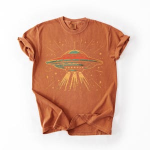 May include: A rust-colored short-sleeve t-shirt featuring a graphic print of a flying saucer in the center. The UFO design includes red, green, and yellow tones with light rays extending downward and small star shapes surrounding the craft.