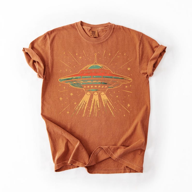 May include: A rust-colored short-sleeve t-shirt featuring a graphic print of a flying saucer in the center. The UFO design includes red, green, and yellow tones with light rays extending downward and small star shapes surrounding the craft.