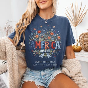 May include: Navy blue t-shirt with the word "AMERICA" in red, white, and blue floral letters. The shirt features a floral wreath design, butterflies, and the text "250TH BIRTHDAY JULY 4, 1776 &ndash; JULY 4, 2026."