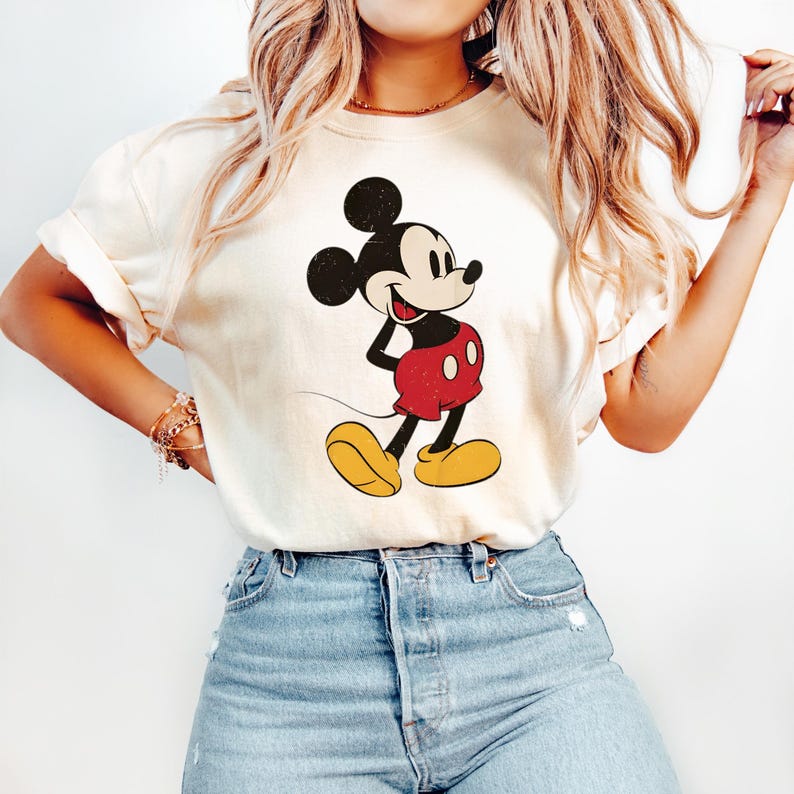 May include: Cream-colored t-shirt featuring a classic Mickey Mouse graphic. The cartoon character is depicted in black, red, and yellow, with a vintage aesthetic. The shirt is paired with blue jeans.