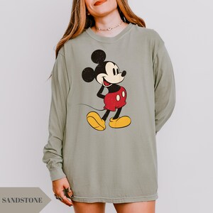 May include: Sage green long-sleeve shirt featuring a distressed graphic of Mickey Mouse in his classic outfit: red shorts, yellow shoes, and black ears. The word "SANDSTONE" is printed at the bottom left.