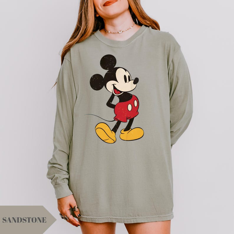 May include: Sage green long-sleeve shirt featuring a distressed graphic of Mickey Mouse in his classic outfit: red shorts, yellow shoes, and black ears. The word "SANDSTONE" is printed at the bottom left.