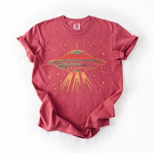 May include: A red short-sleeved t-shirt featuring a graphic print of a flying saucer emitting a beam of light. The design includes yellow stars and lines radiating from the spacecraft. The shirt has a crew neckline and a visible brand label at the neck.