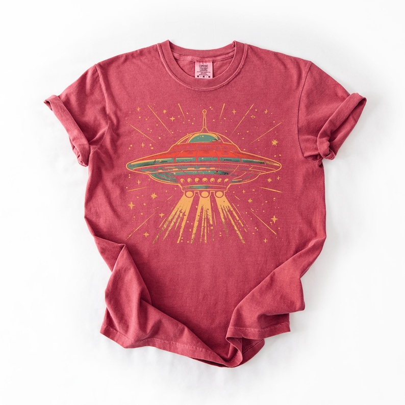 May include: A red short-sleeved t-shirt featuring a graphic print of a flying saucer emitting a beam of light. The design includes yellow stars and lines radiating from the spacecraft. The shirt has a crew neckline and a visible brand label at the neck.