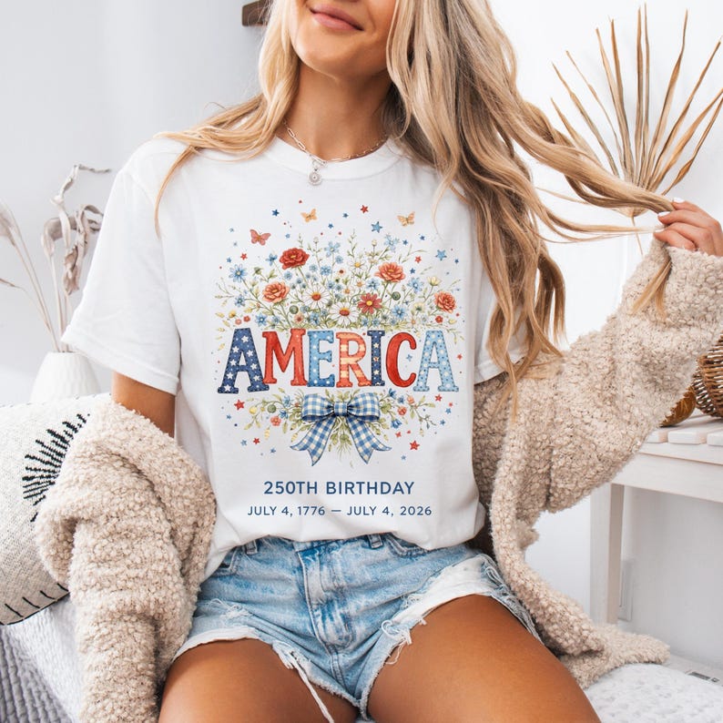 May include: White t-shirt with a floral design and the word "AMERICA" in red, white, and blue. The design includes a blue and white bow, flowers, and butterflies. Text below reads "250th BIRTHDAY JULY 4, 1776 - JULY 4, 2026."