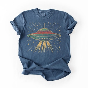 May include: A blue short-sleeve t-shirt featuring a graphic of a flying saucer emitting a yellow light beam. The saucer is depicted in shades of green and red with a star-filled background. The design has a distressed, textured appearance.