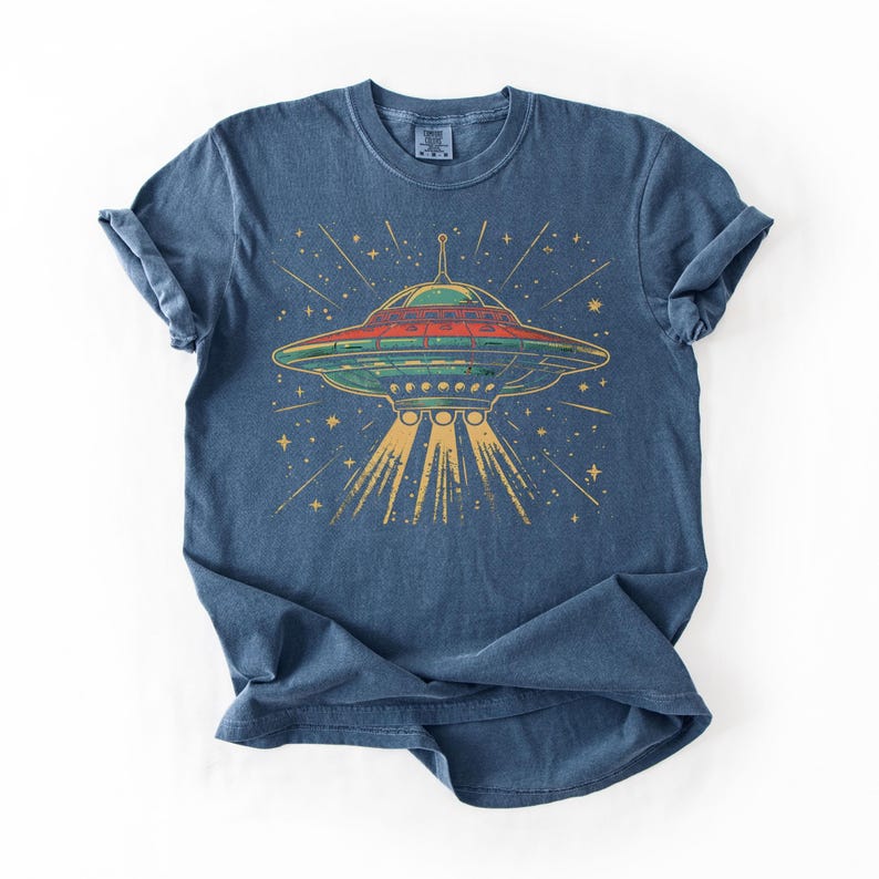 May include: A blue short-sleeve t-shirt featuring a graphic of a flying saucer emitting a yellow light beam. The saucer is depicted in shades of green and red with a star-filled background. The design has a distressed, textured appearance.