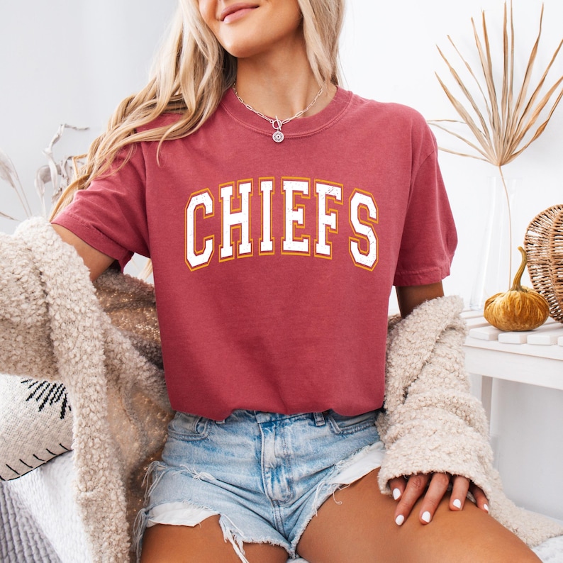Op de afbeelding: Een T-shirt met korte mouwen en een ronde hals in een gedempte rode kleur. Op de voorkant staat het woord CHIEFS in een groot, gebogen, wit lettertype in collegestijl met een dunne gele omlijning. De stof heeft een vintage textuur.