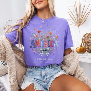 May include: A lilac-colored t-shirt with the word "AMERICA" in a decorative font, surrounded by floral and butterfly graphics. A blue and white plaid bow is below the word. The text "250TH BIRTHDAY JULY 4, 1776 - JULY 4, 2026" is printed below the bow.