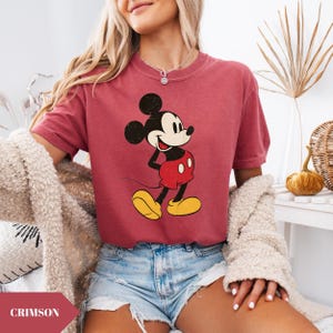 May include: Crimson-colored t-shirt featuring a classic Mickey Mouse graphic. The cartoon character is depicted in black, red, yellow, and white. The shirt has short sleeves and a crew neck. The word "CRIMSON" is in a banner at the bottom left.