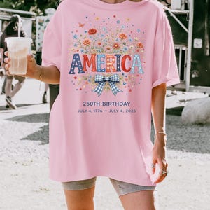 May include: Pink t-shirt with the word "AMERICA" in patriotic colors, surrounded by floral and butterfly graphics. The shirt also features the text "250TH BIRTHDAY JULY 4, 1776 - JULY 4, 2026".