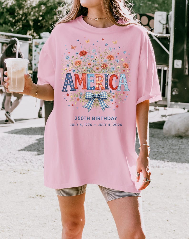 May include: Pink t-shirt with the word "AMERICA" in patriotic colors, surrounded by floral and butterfly graphics. The shirt also features the text "250TH BIRTHDAY JULY 4, 1776 - JULY 4, 2026".