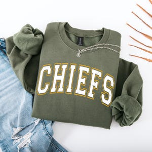 May include: Olive green sweatshirt with the word "CHIEFS" in white block letters with a gold outline. A silver necklace with a circular pendant rests on the sweatshirt. The sweatshirt is paired with blue jeans.