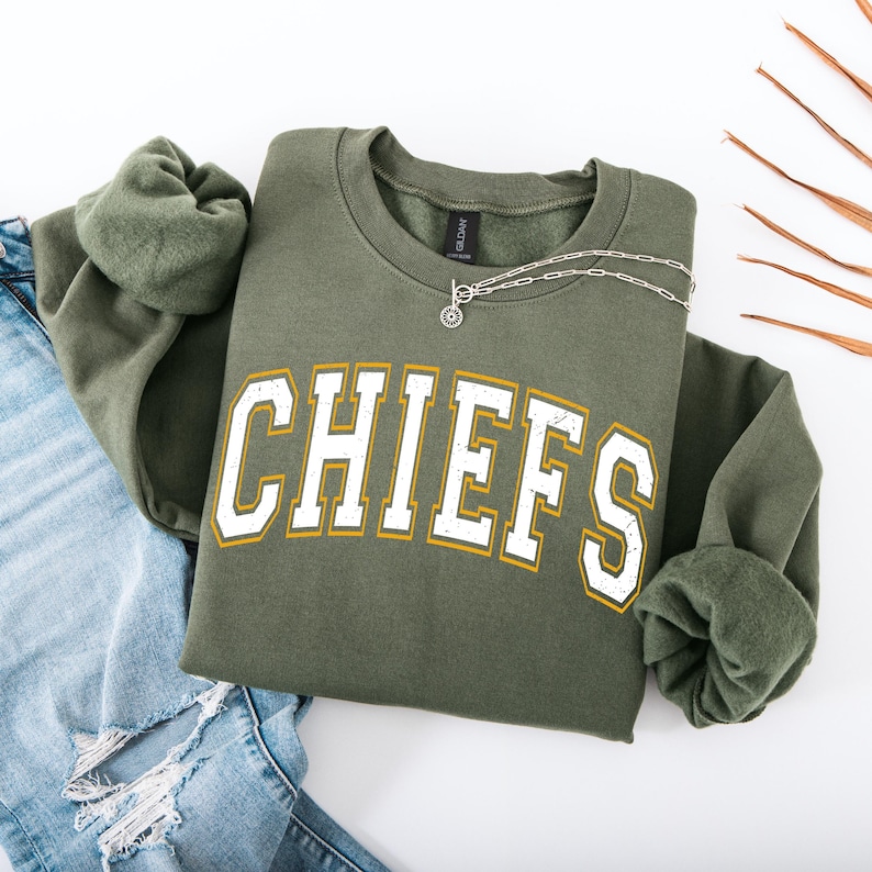 May include: Olive green sweatshirt with the word "CHIEFS" in white block letters with a gold outline. A silver necklace with a circular pendant rests on the sweatshirt. The sweatshirt is paired with blue jeans.