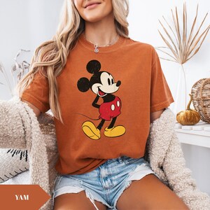 May include: Burnt orange t-shirt featuring a vintage-style Mickey Mouse graphic. Mickey is depicted in his classic pose, wearing red shorts and yellow shoes. The shirt has a relaxed fit and short sleeves, perfect for casual wear.