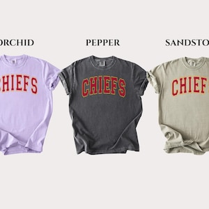 Op de afbeelding: Drie t-shirts met korte mouwen in de kleuren orchidee, peper en zandsteen. Elk shirt heeft het woord CHIEFS in een rood gebogen lettertype met een gele omlijning op de borst gedrukt. De shirts worden naast elkaar getoond met labels erboven.