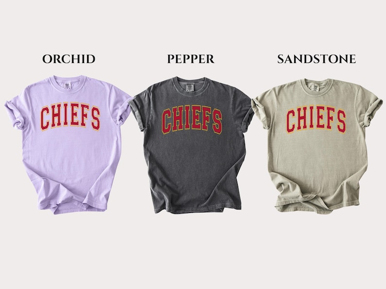 Op de afbeelding: Drie t-shirts met korte mouwen in de kleuren orchidee, peper en zandsteen. Elk shirt heeft het woord CHIEFS in een rood gebogen lettertype met een gele omlijning op de borst gedrukt. De shirts worden naast elkaar getoond met labels erboven.