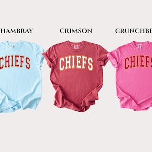 Op de afbeelding: Drie T-shirts met korte mouwen en ronde hals in lichtblauw, karmozijnrood en roze. Elk shirt heeft het woord CHIEFS in een wit gebogen lettertype met een rode omlijning op de borst. De kleuren staan bovenaan vermeld als CHAMBRAY, CRIMSON en CRUNCHBERRY.