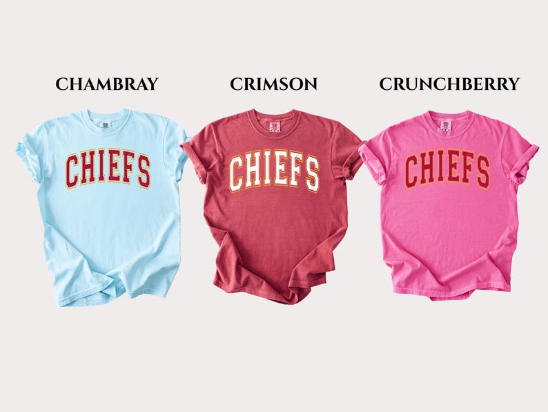 Op de afbeelding: Drie T-shirts met korte mouwen en ronde hals in lichtblauw, karmozijnrood en roze. Elk shirt heeft het woord CHIEFS in een wit gebogen lettertype met een rode omlijning op de borst. De kleuren staan bovenaan vermeld als CHAMBRAY, CRIMSON en CRUNCHBERRY.