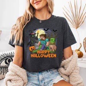 May include: Dark grey t-shirt with a Halloween graphic. The design features pixelated characters, a witch, a pumpkin, and the text "HAPPY HALLOWEEN" in orange. Bats and a full moon are also visible.