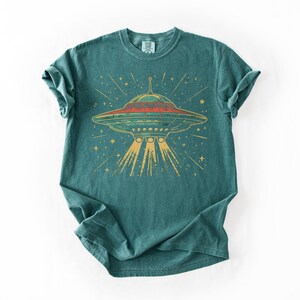 May include: A teal short-sleeve t-shirt featuring a graphic print of a flying saucer emitting a yellow light beam. The saucer is depicted in shades of red, green, and yellow, surrounded by small yellow stars against the dark teal fabric background.