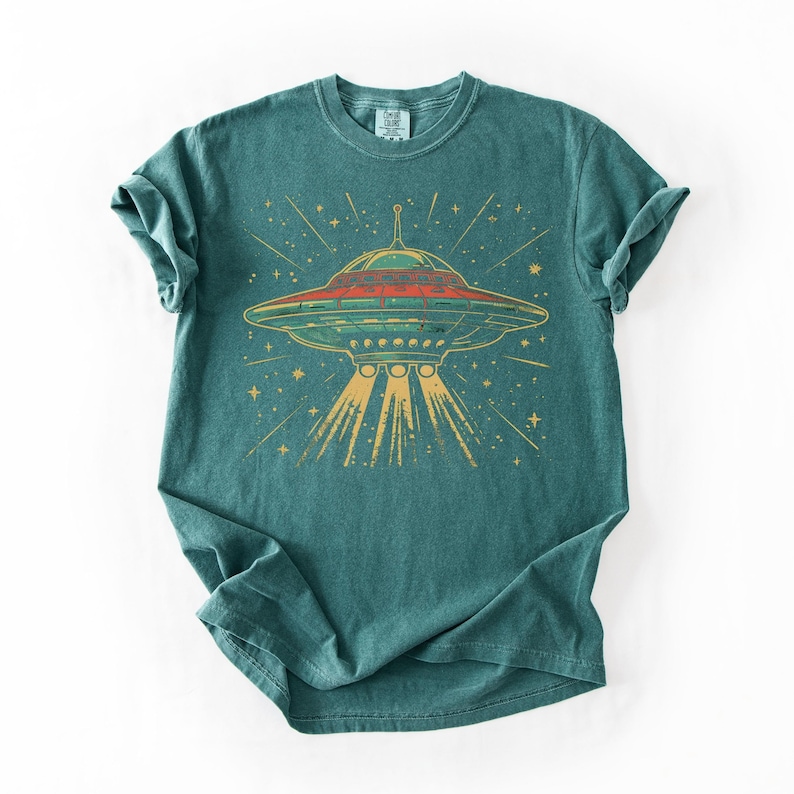 May include: A teal short-sleeve t-shirt featuring a graphic print of a flying saucer emitting a yellow light beam. The saucer is depicted in shades of red, green, and yellow, surrounded by small yellow stars against the dark teal fabric background.