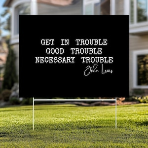 Könnte beinhalten: Schwarzes Gartenschild mit weißem Text: „GET IN TROUBLE GOOD TROUBLE NECESSARY TROUBLE“ und der Unterschrift „John Lewis“. Das Schild wird auf einem grünen Rasen mit weißem Metallrahmen angezeigt.