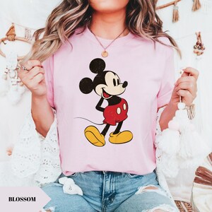 May include: A light pink t-shirt featuring a distressed graphic of Mickey Mouse in his classic pose. Mickey is depicted in black, red, yellow, and white. The word "BLOSSOM" is printed in a white font on a light pink shape.