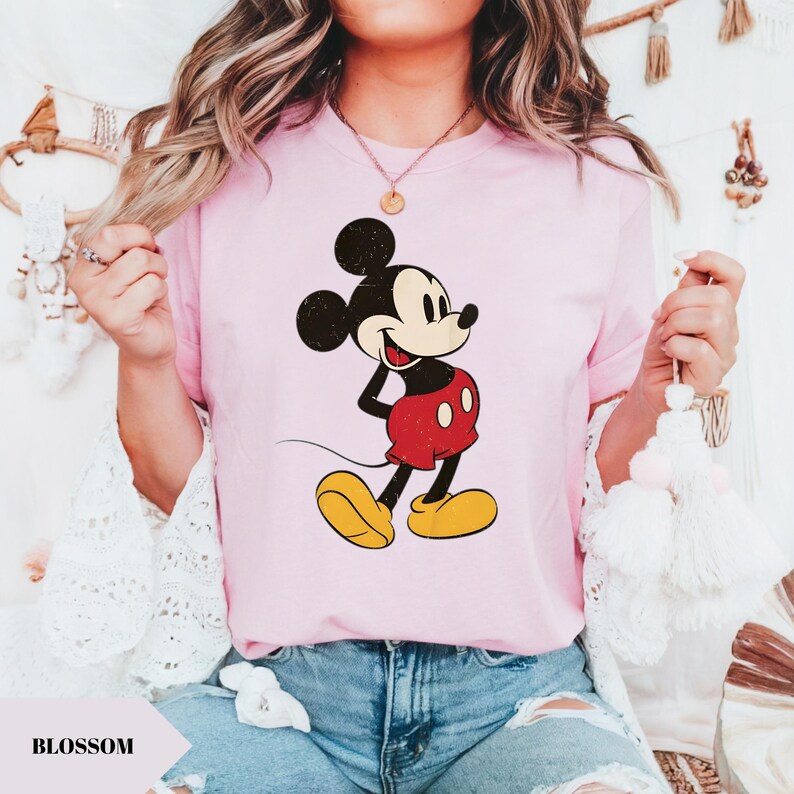 May include: A light pink t-shirt featuring a distressed graphic of Mickey Mouse in his classic pose. Mickey is depicted in black, red, yellow, and white. The word "BLOSSOM" is printed in a white font on a light pink shape.