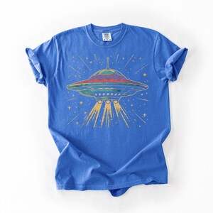 May include: A bright blue short-sleeve t-shirt featuring a graphic print of a flying saucer in shades of red, green, and yellow. The design includes a central UFO emitting three light beams downward, surrounded by small yellow stars and radiating lines.