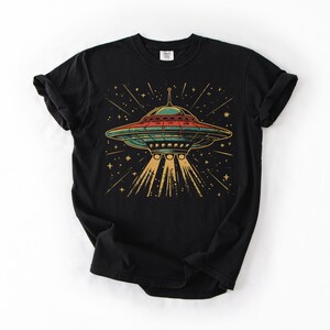 May include: A black short-sleeve t-shirt featuring a graphic print of a retro-style flying saucer emitting a bright yellow light beam. The saucer is colored in shades of teal and red, surrounded by a field of yellow stars against the black fabric.