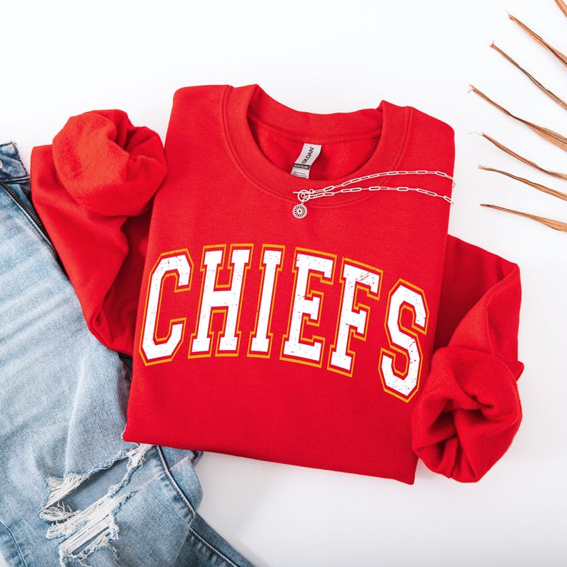 May include: A red crewneck sweatshirt with the word "CHIEFS" in white block letters with yellow trim. A silver necklace with a pendant is draped over the sweatshirt. The sweatshirt is paired with blue jeans.